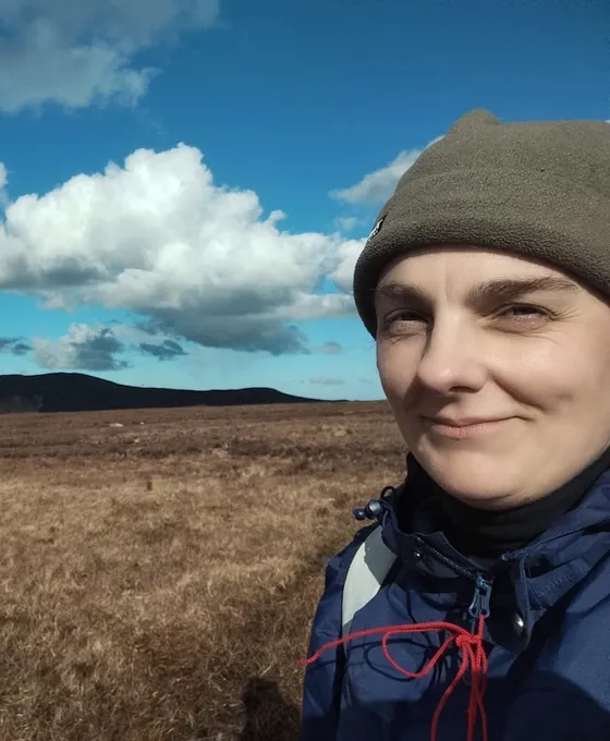 Orna Ní Choileáin ag siúl ar an sliabh faoi spéir ghorm agus scamaill bhána.